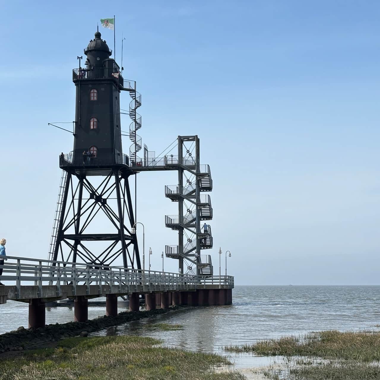 Ein hoher, schwarzer Leuchtturm auf Stelzen mit einer Wendeltreppe, verbunden mit einem Steg über ruhigem Wasser unter einem klaren blauen Himmel.