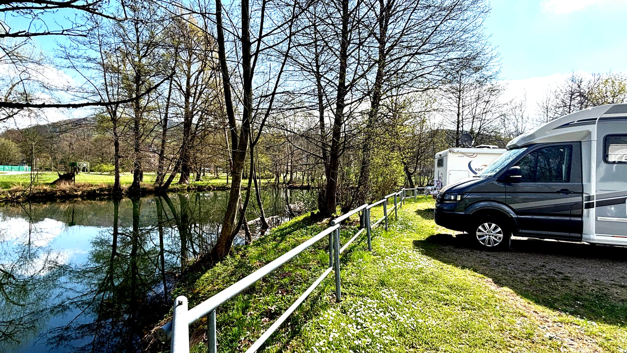 Wohnmobile, die an einem ruhigen Fluss geparkt sind, umgeben von Bäumen und Grün unter einem klaren blauen Himmel.