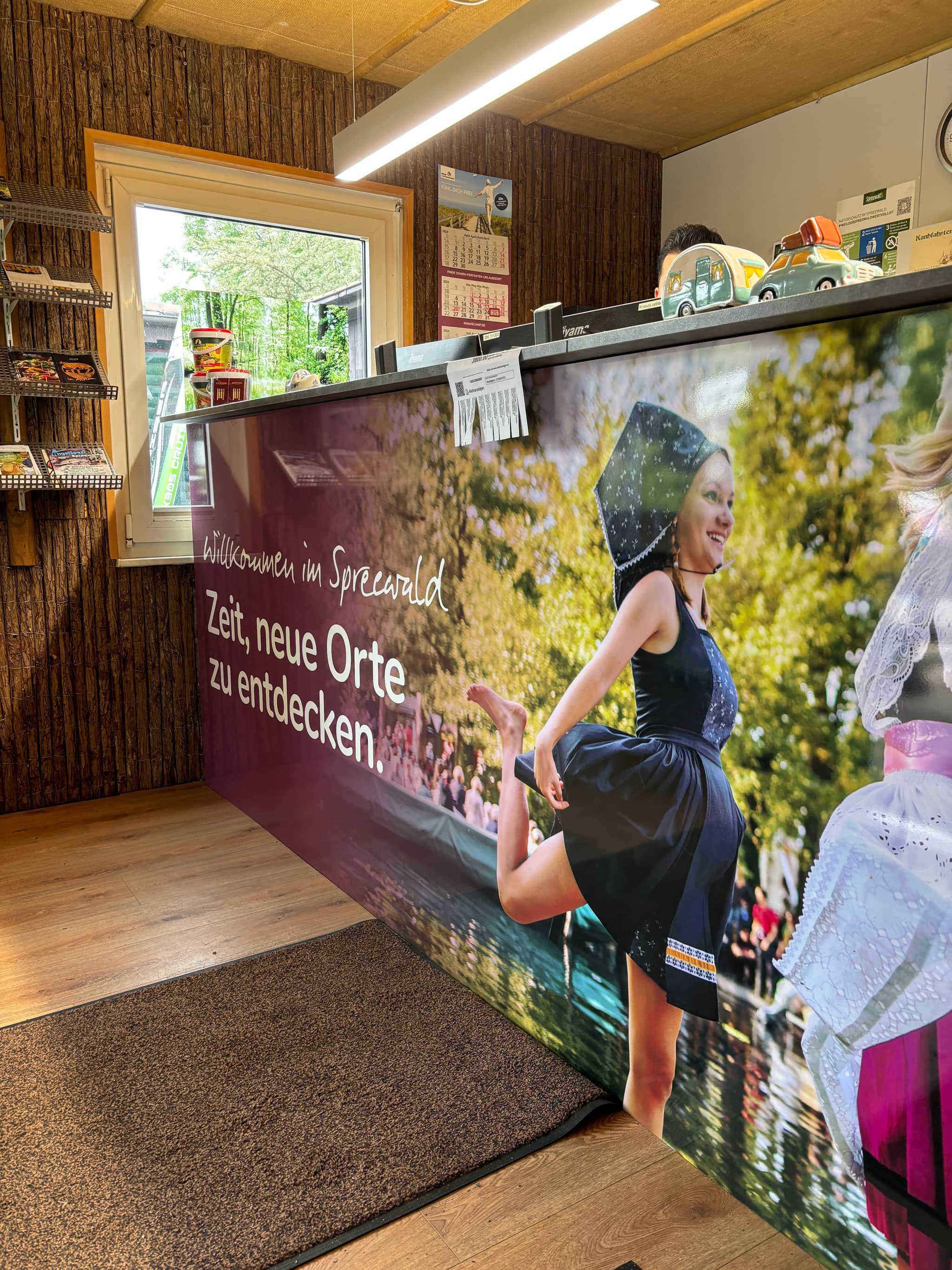 Empfangsbereich mit einem großen Foto einer Frau in traditioneller Kleidung, Text in deutscher Sprache, Holzwänden und einem Fenster mit Blick ins Grüne.
