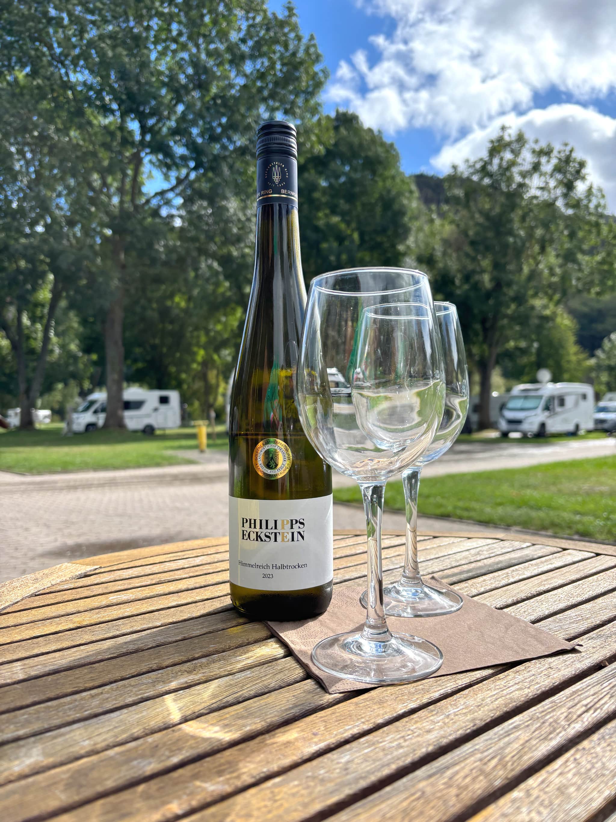 Eine Flasche Weißwein und zwei leere Gläser auf einem Holztisch im Freien, mit Bäumen und geparkten Wohnmobilen im Hintergrund unter einem teilweise bewölkten Himmel.