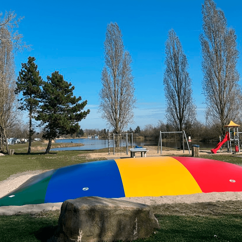 Colorful inflatable play structure in a park with tall trees, a swing set, and a slide. A lake is visible in the background under a clear blue sky.