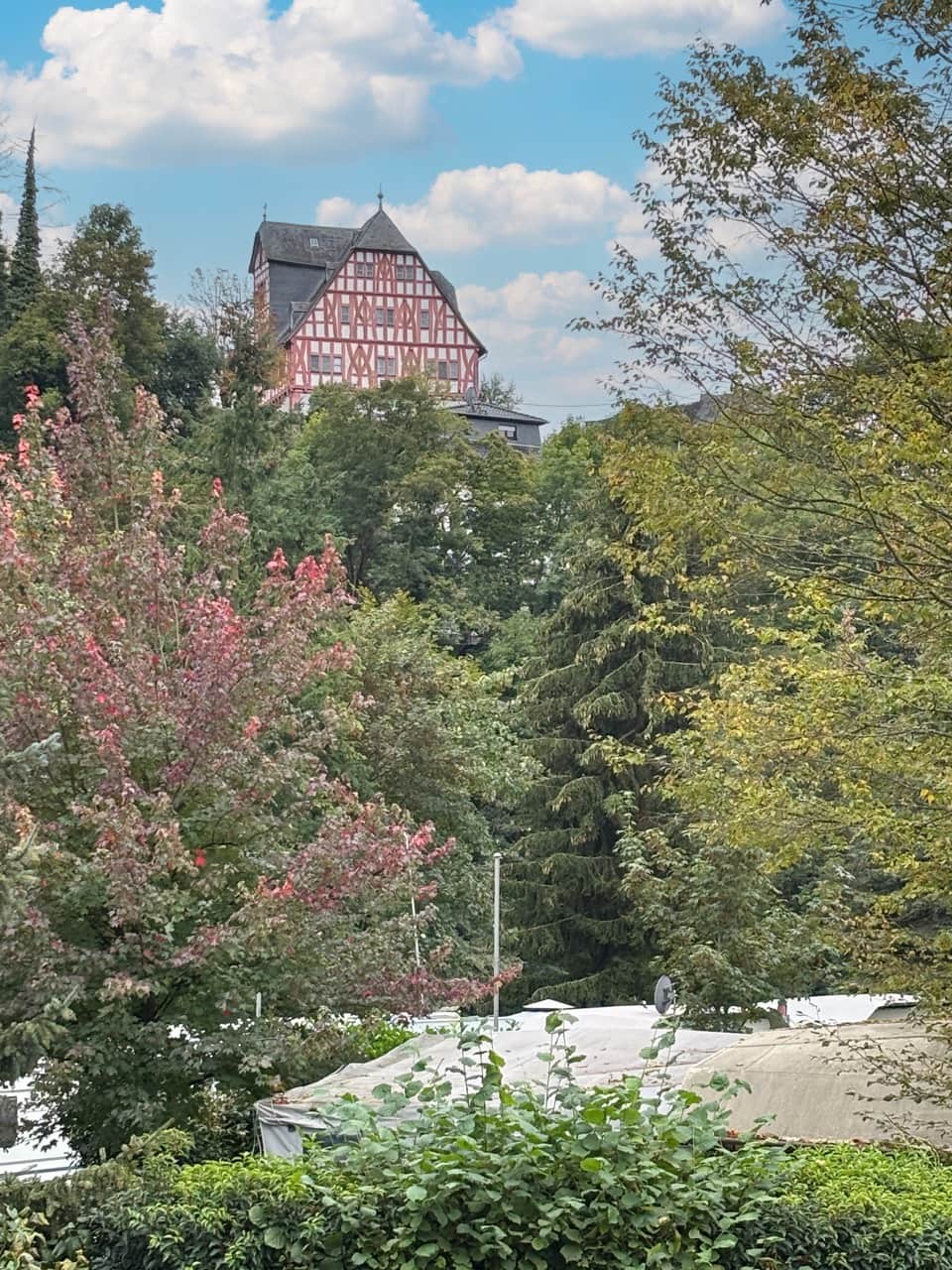 Ein traditionelles Fachwerkhaus mit steilem Dach ist teilweise durch üppiges grünes und rotes Laub unter einem blauen Wolkenhimmel sichtbar.