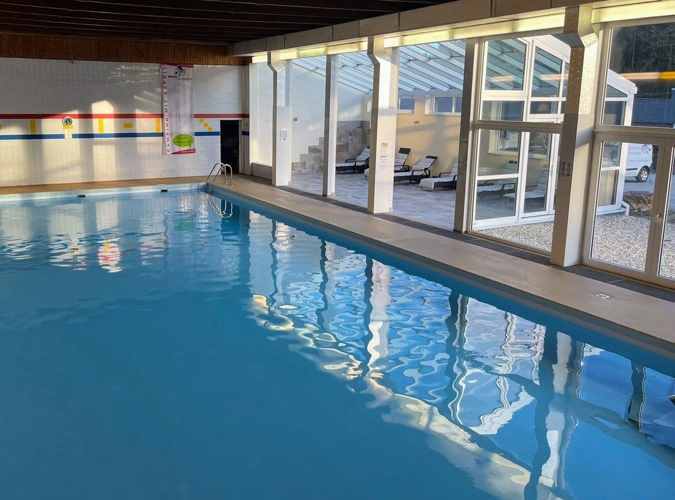 Innenschwimmbad mit ruhigem, blauem Wasser, großen Fenstern auf der rechten Seite und Liegestühlen draußen in einem sonnigen Bereich.