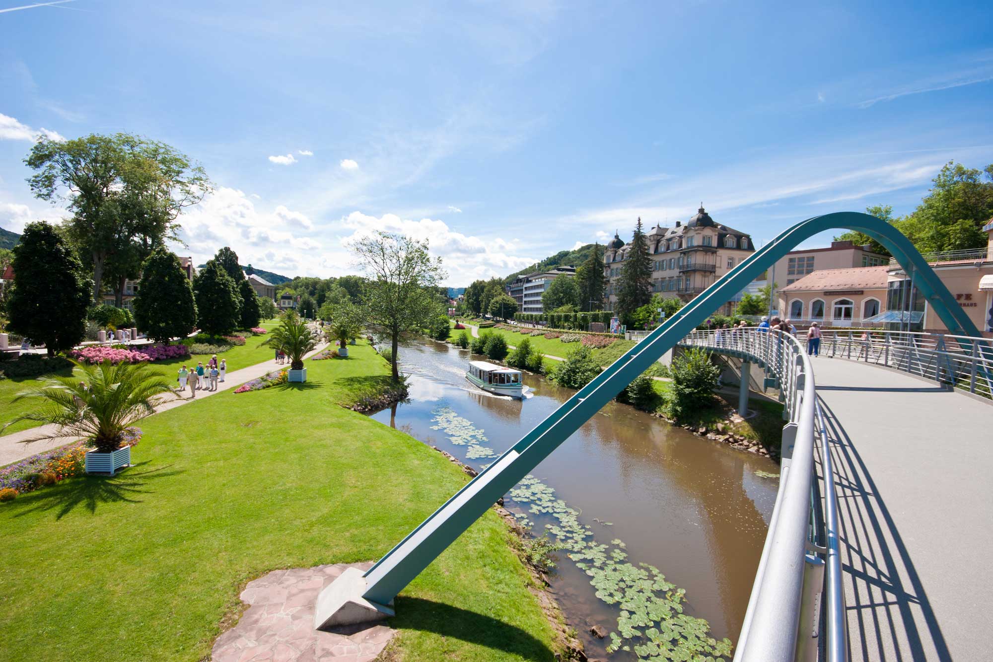 Ein malerischer Blick auf einen Park mit einer modernen Brücke über einen Fluss, umgeben von Grünflächen, Bäumen und historischen Gebäuden unter einem klaren blauen Himmel.
