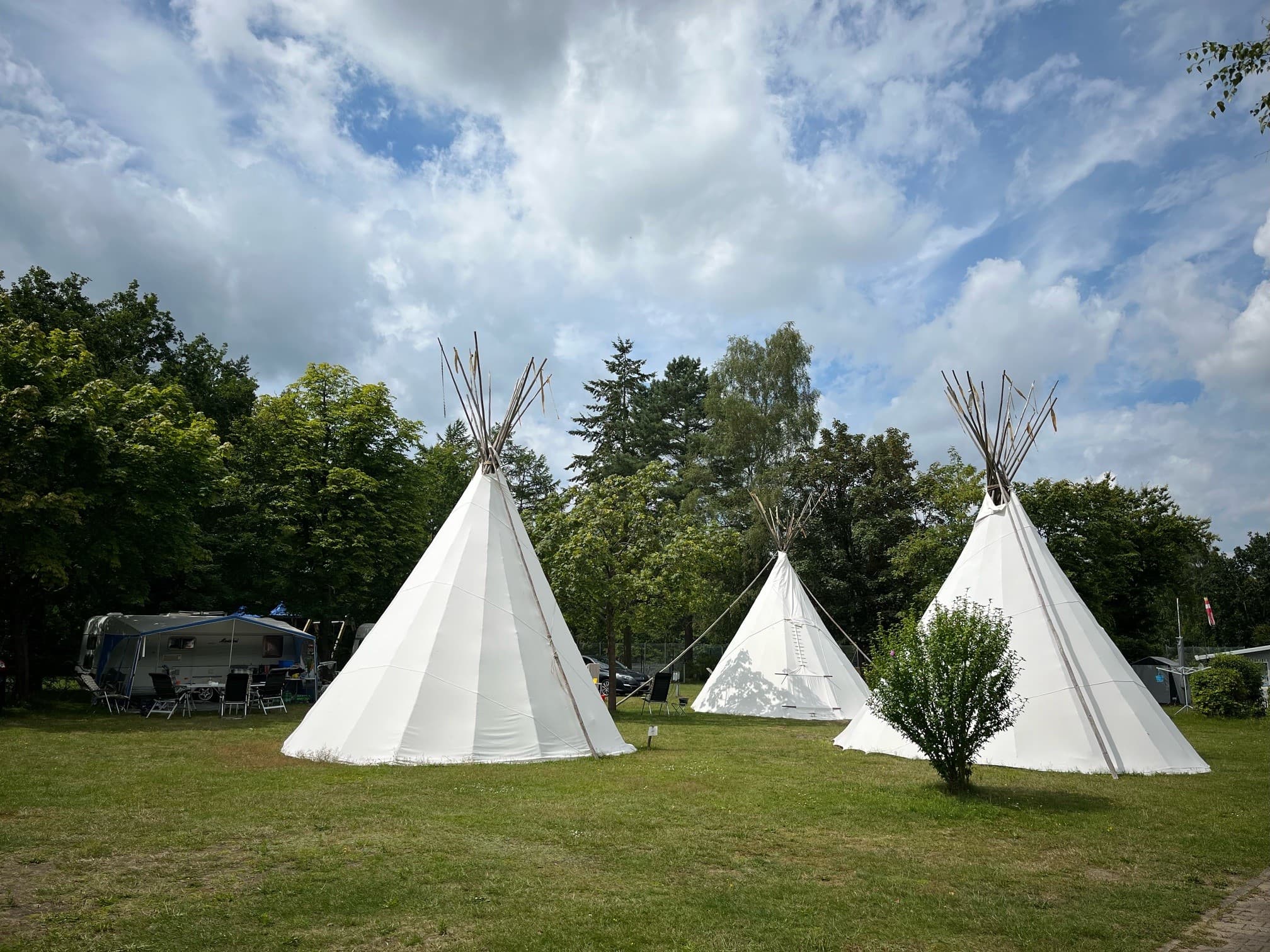 Drei weiße Tipis auf einer von Bäumen umgebenen Wiese unter einem teilweise bewölkten Himmel, mit einem Wohnwagen und Stühlen in der Nähe.