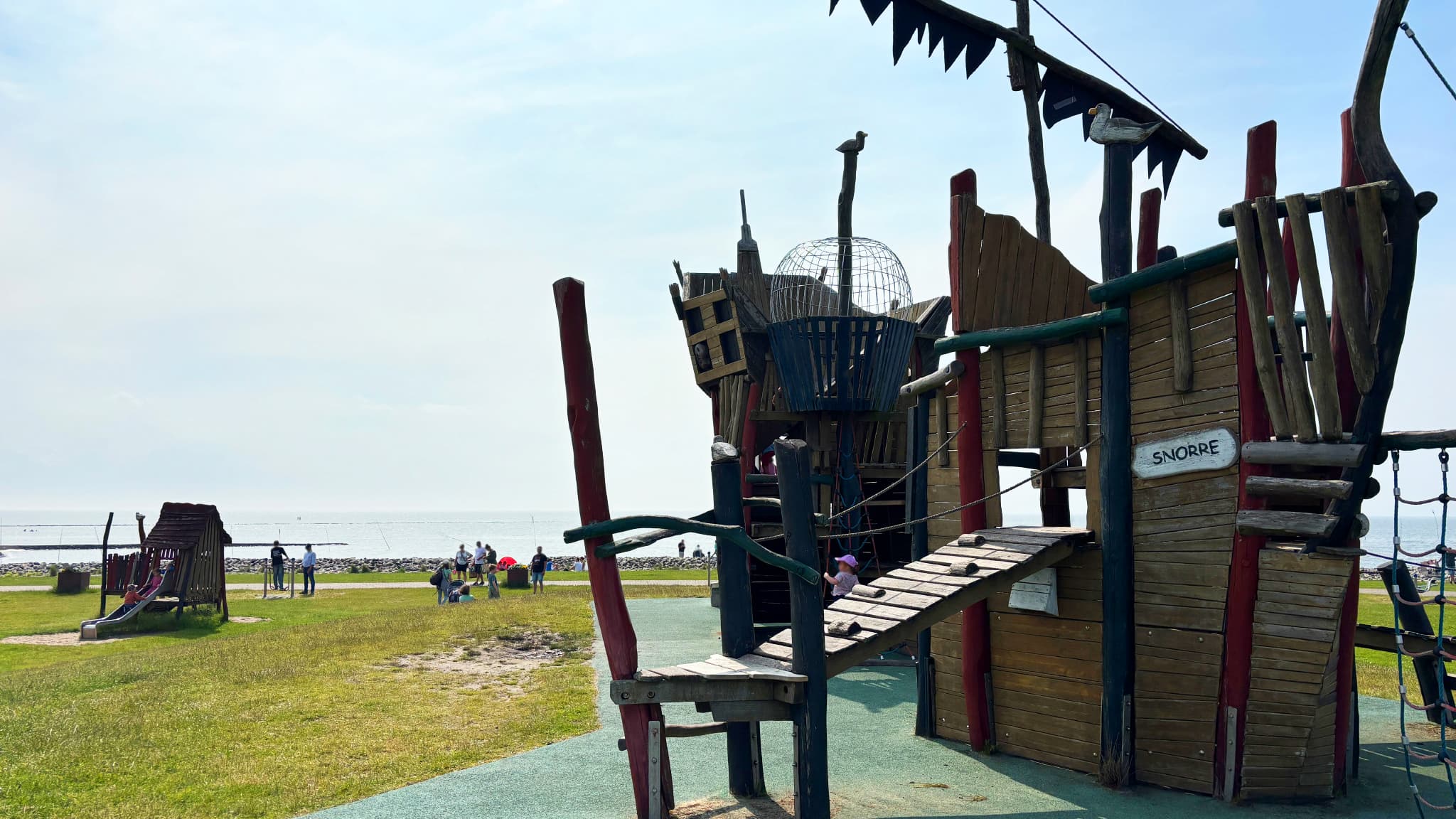 Spielplatz mit einem hölzernen Piratenschiff Struktur auf Gras in der Nähe eines Strandes, mit Menschen im Hintergrund unter einem bewölkten Himmel.