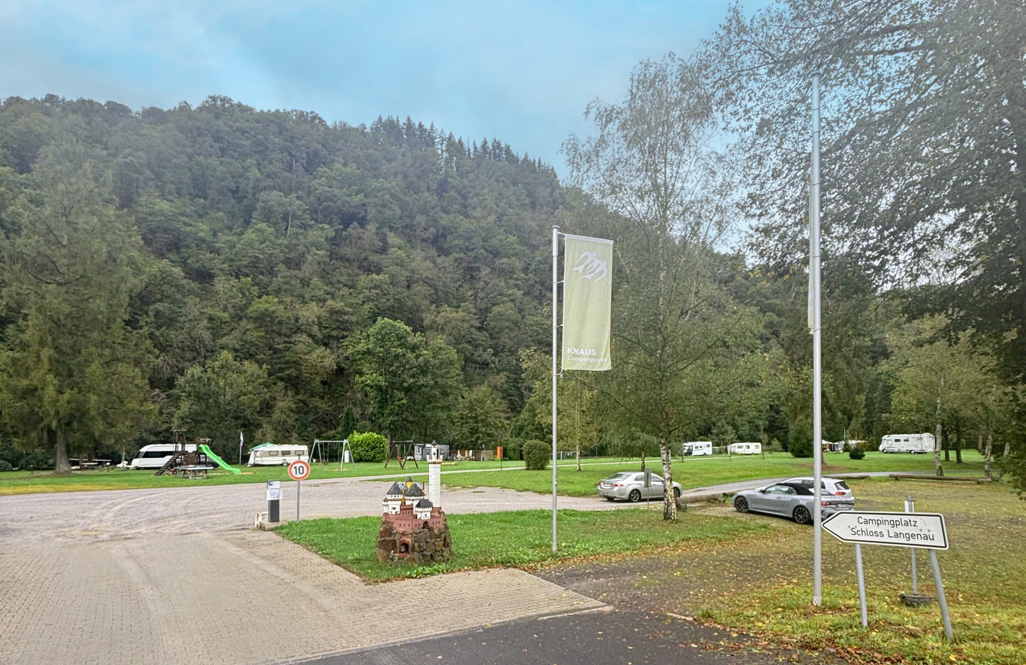 Campingplatz mit geparkten Wohnwagen, Autos und einem Spielplatz. Umgeben von üppigen grünen Bäumen und Hügeln unter einem bewölkten Himmel. Schilder am Eingang sichtbar.
