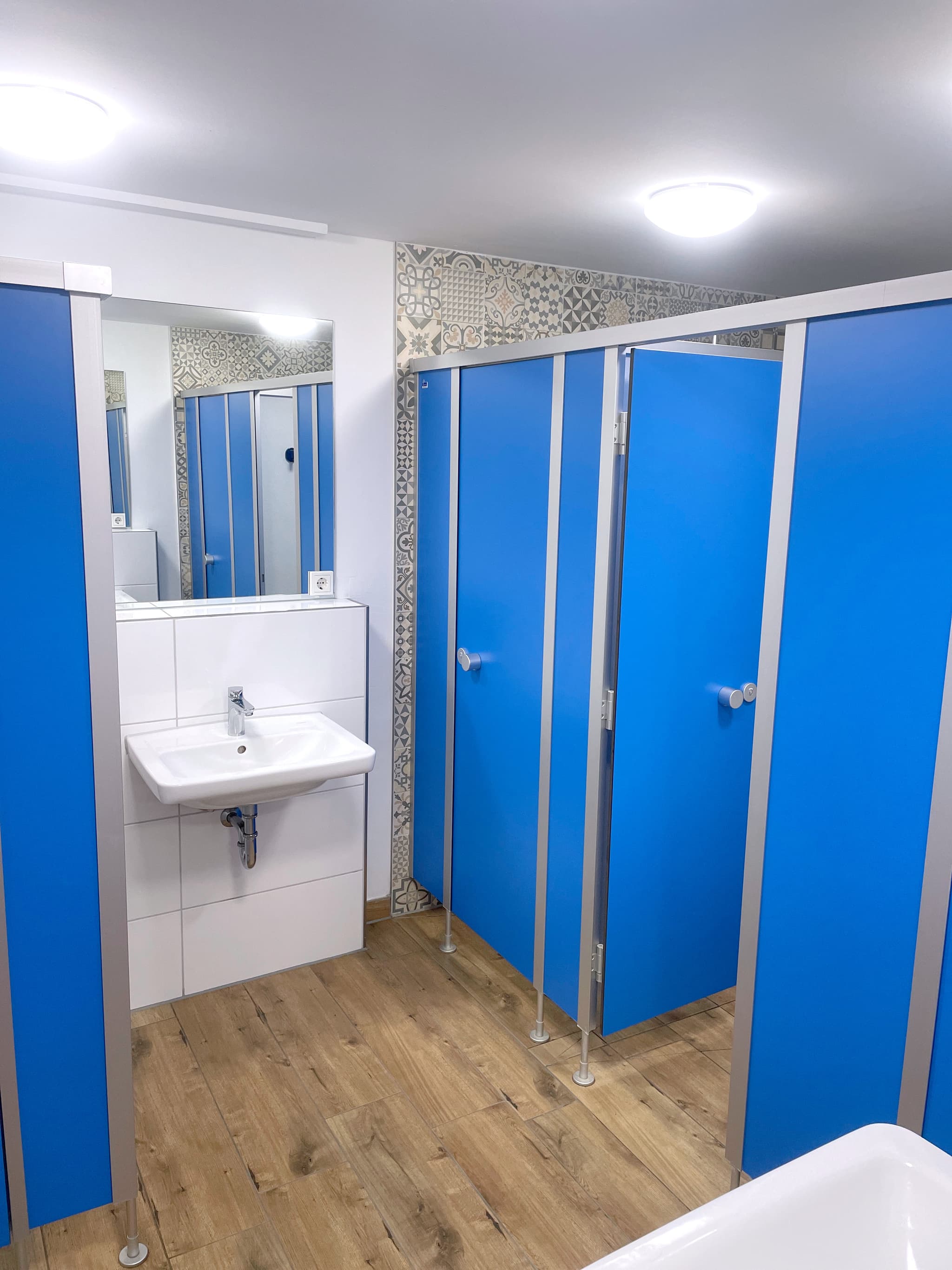 Eine  Toilette mit blauen Kabinentüren, einem weißen Waschbecken, einem Spiegel und Holzfußboden.