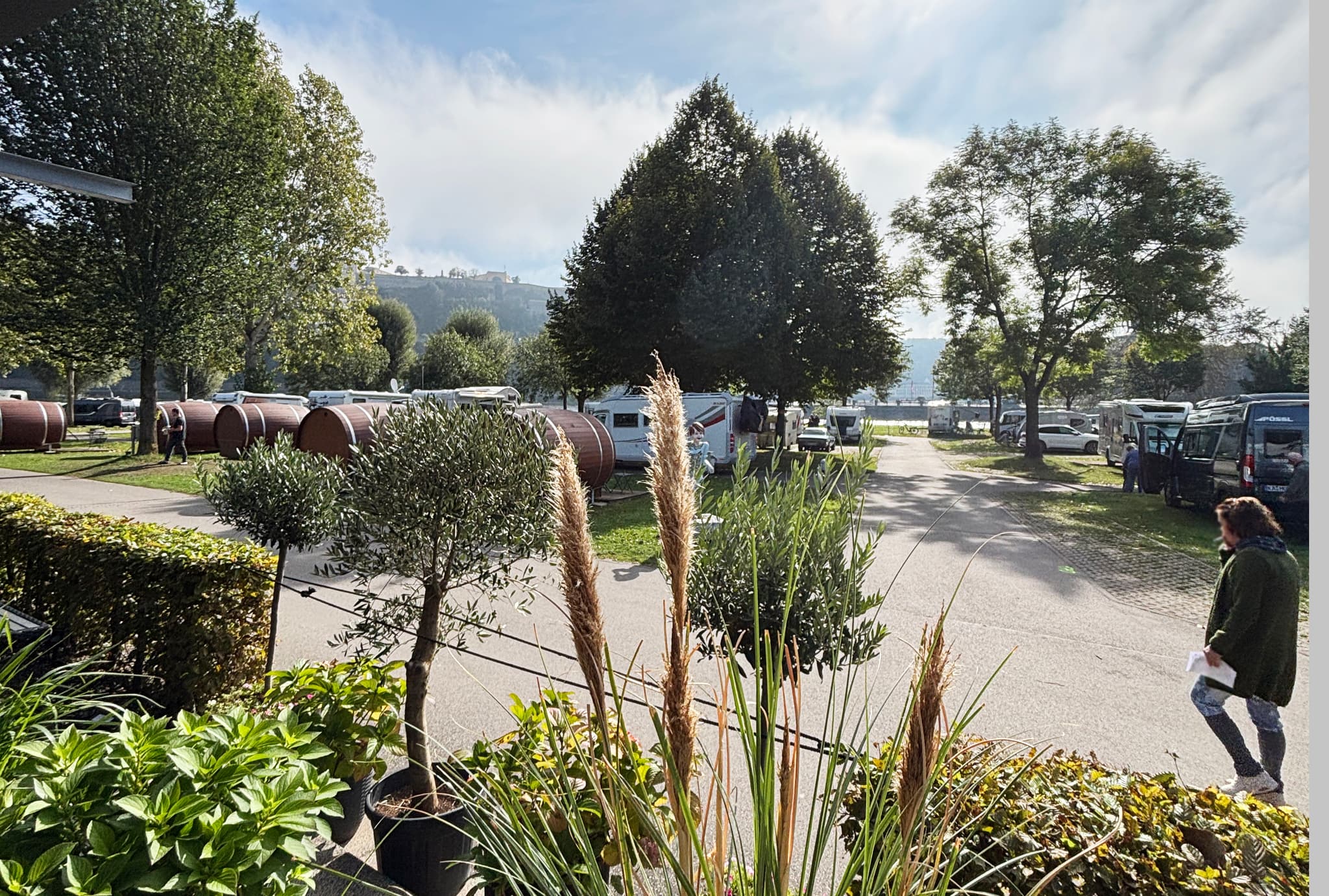 Ein malerischer Campingplatz mit Bäumen, geparkten Wohnmobilen und einer Person, die unter einem teilweise bewölkten Himmel einen Weg entlangläuft.