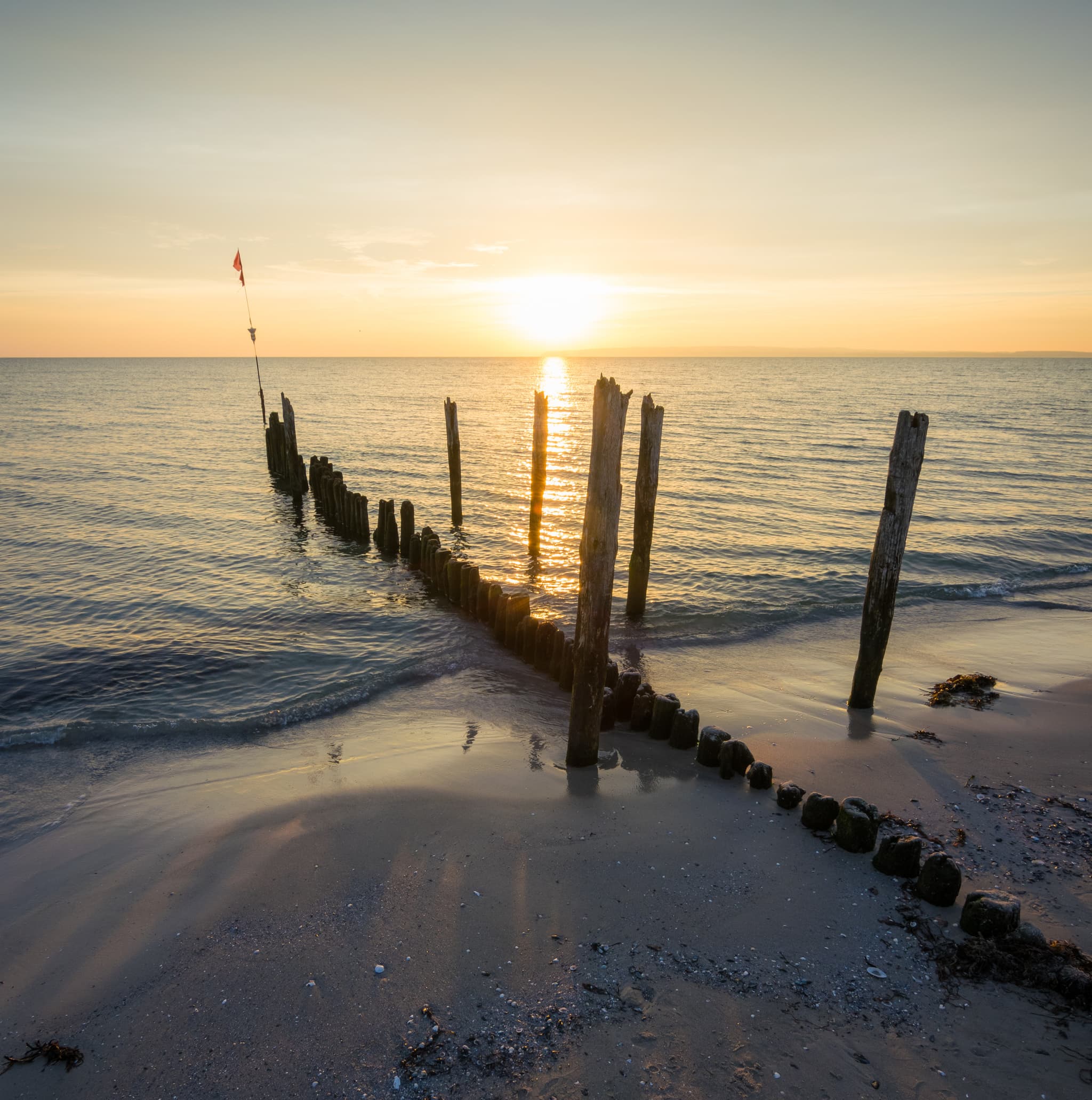 Sonnenuntergang über einer ruhigen See mit verwitterten Holzpfählen, die von einem Sandstrand ins Wasser ragen und lange Schatten werfen.