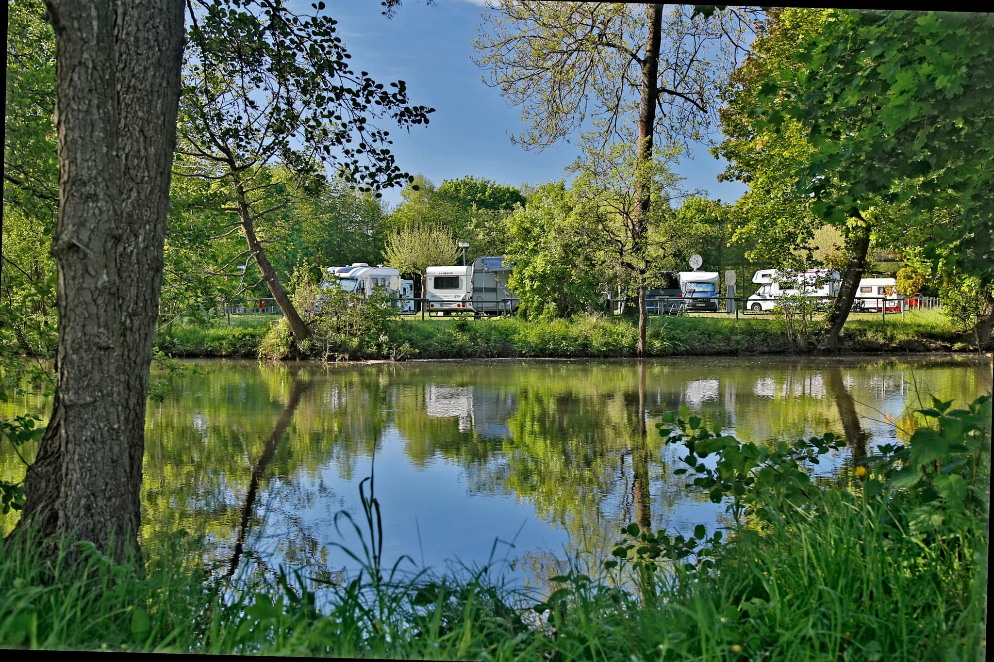 Eine ruhige Flussszene mit Bäumen und Gras, die sich im Wasser spiegeln. Im Hintergrund sind mehrere geparkte Wohnmobile bei klarem Himmel zu sehen.