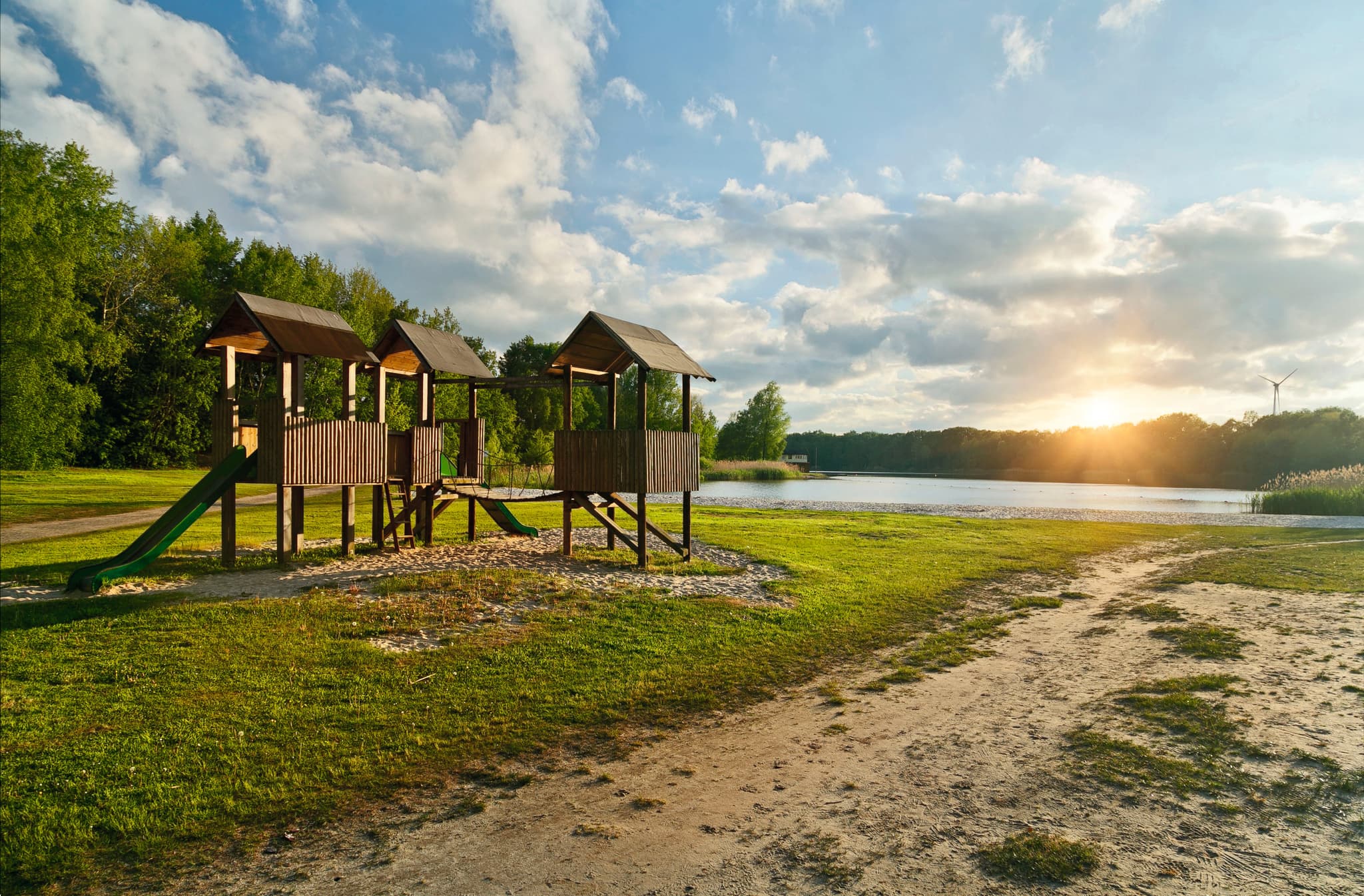 Spielplatz mit Holzkonstruktionen in der Nähe eines ruhigen Sees bei Sonnenuntergang, umgeben von üppigem Grün und einem sandigen Weg.