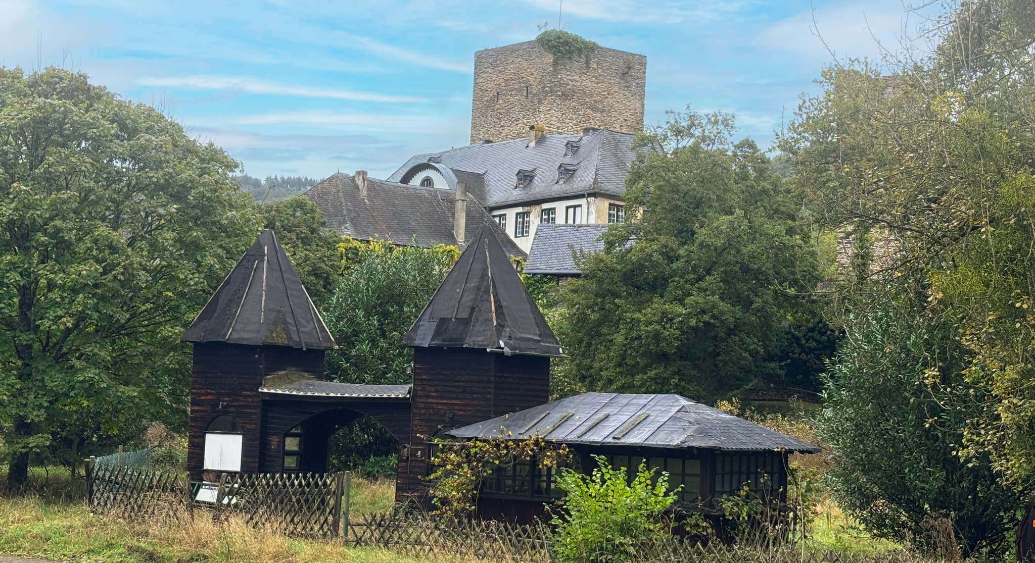 Eine rustikale Holzkonstruktion mit spitzen Dächern in einer üppigen Landschaft, mit einem Steinturm und einem Gebäude, die teilweise im Hintergrund sichtbar sind.