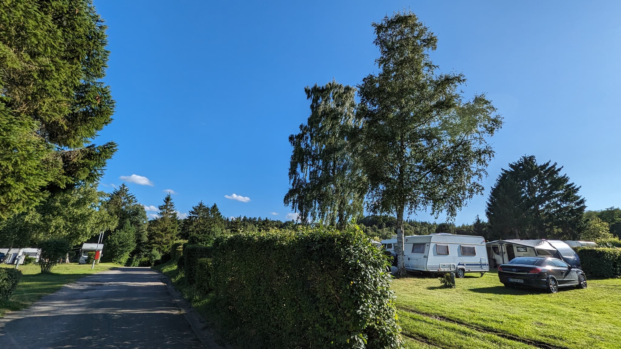 Ein sonniger Campingplatz mit Wohnwagen, einem Auto und Bäumen unter einem strahlend blauen Himmel. Eine schmale Straße führt entlang gestutzter Hecken.
