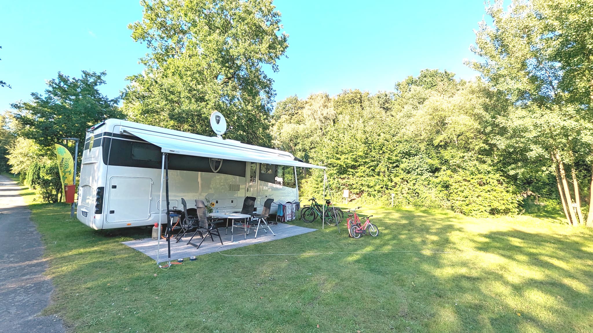 Ein geparktes Wohnmobil mit ausgefahrener Markise, umgeben von Stühlen, einem Tisch und Fahrrädern auf einer Wiese mit Bäumen im Hintergrund.
