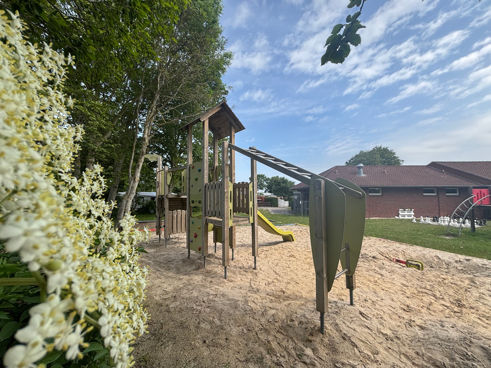 Spielplatz mit einem Klettergerüst und einer Rutsche auf sandigem Boden, umgeben von Bäumen und einem Gebäude im Hintergrund unter einem teilweise bewölkten Himmel.