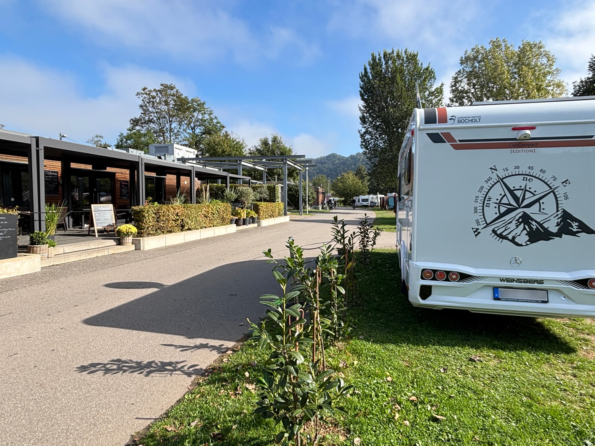 Ein Wohnmobil steht in der Nähe eines modernen Gebäudes mit Sitzgelegenheiten im Freien, umgeben von Grünflächen und Bäumen unter einem teilweise bewölkten Himmel.