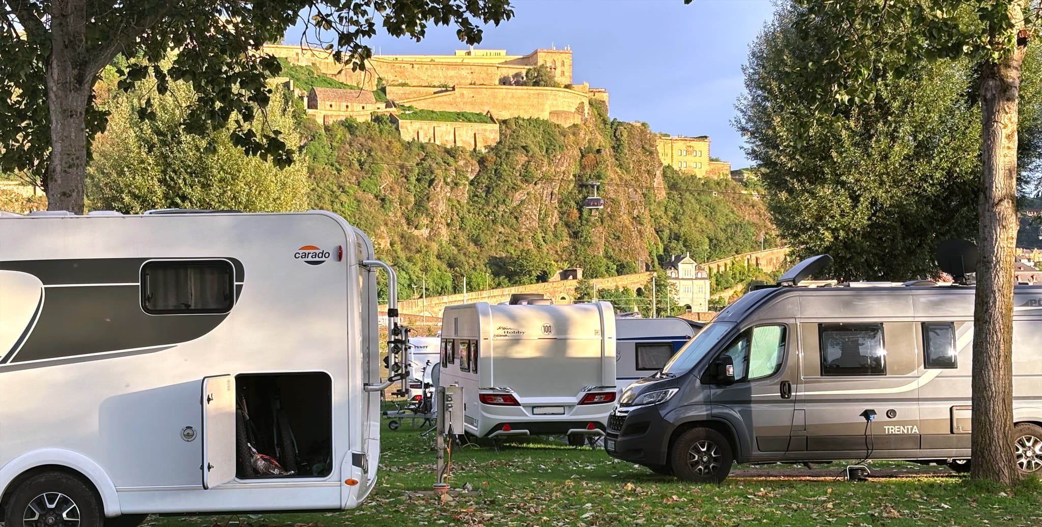 Auf einer Wiese mit Bäumen geparkte Wohnmobile unter einem sonnigen Himmel. Eine historische Festung ist auf einem Hügel im Hintergrund zu sehen.