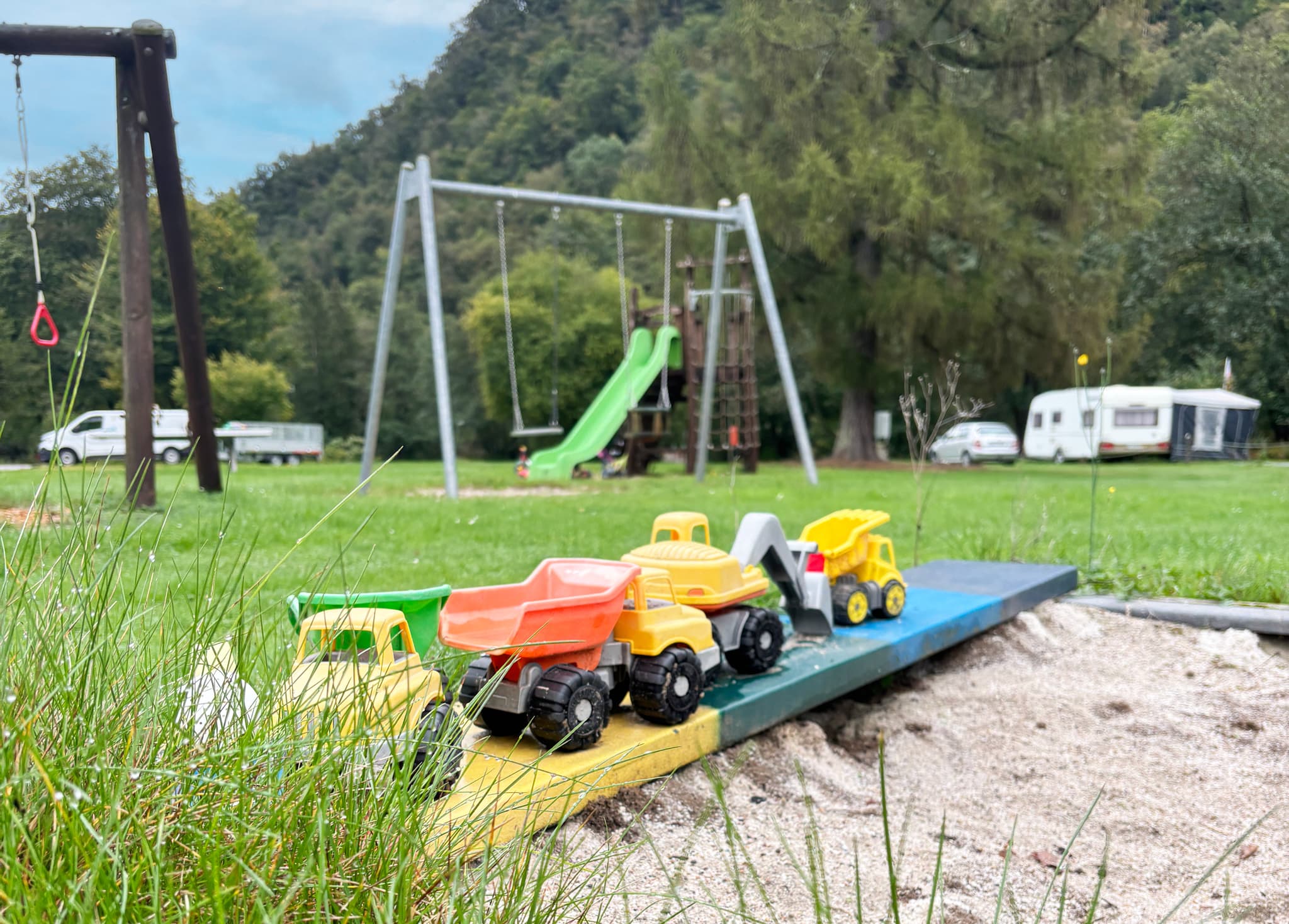 Spielzeugautos auf einer Sandkastenbrücke auf einem Spielplatz mit einer Schaukel, einer Rutsche, einer Wiese und geparkten Wohnwagen im Hintergrund.