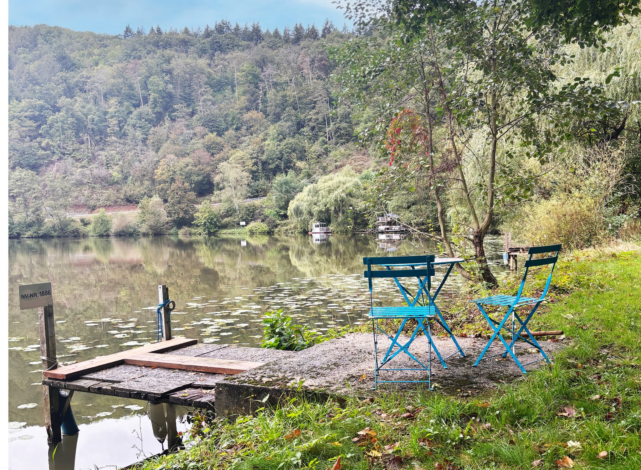 Zwei blaue Stühle und ein Tisch auf einer Terrasse am See, umgeben von Bäumen und Grün, mit einem kleinen Steg, der ins Wasser hineinragt.