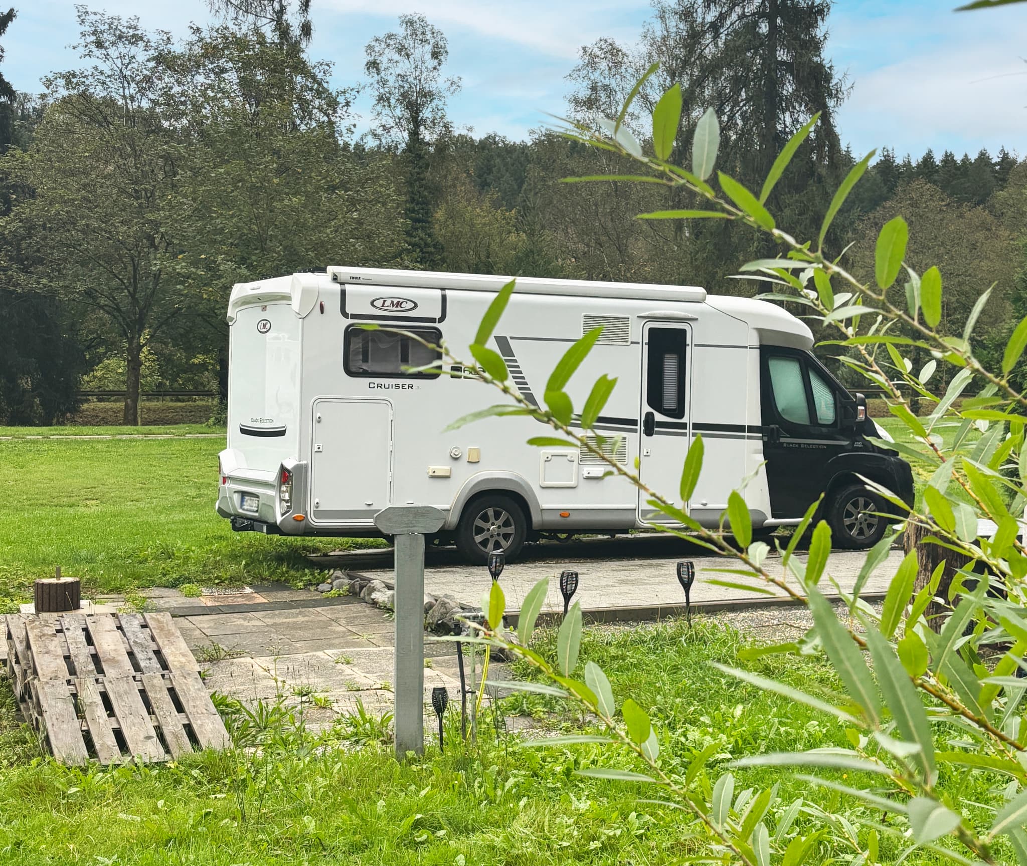 Ein weißes Wohnmobil, geparkt auf einer Wiese mit Bäumen im Hintergrund, umgeben von üppigem Grün und einem klaren blauen Himmel.