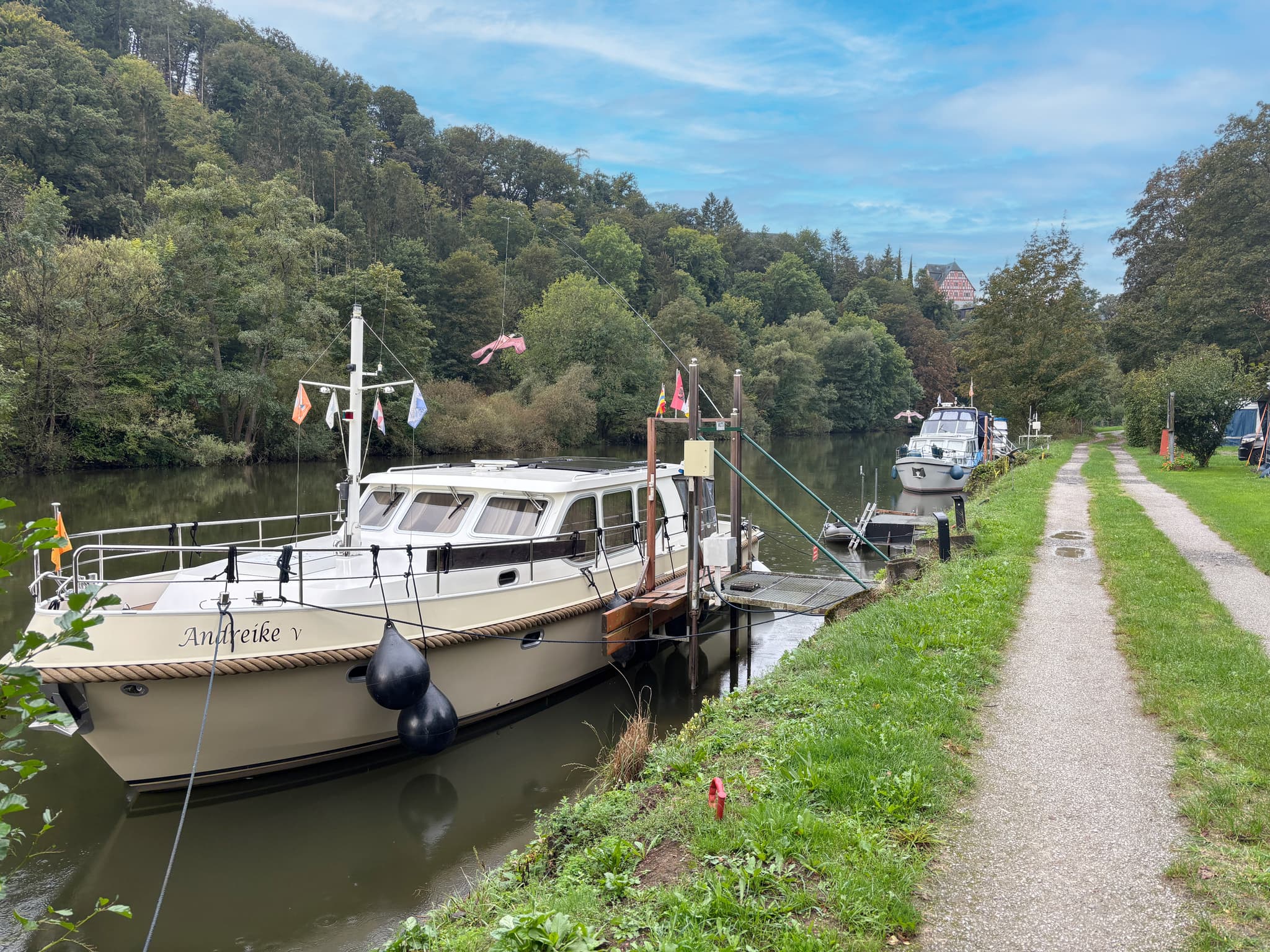 Boote, die an einem ruhigen Fluss mit einem grasbewachsenen Weg und einer bewaldeten Fläche angedockt sind, unter einem teilweise bewölkten Himmel.