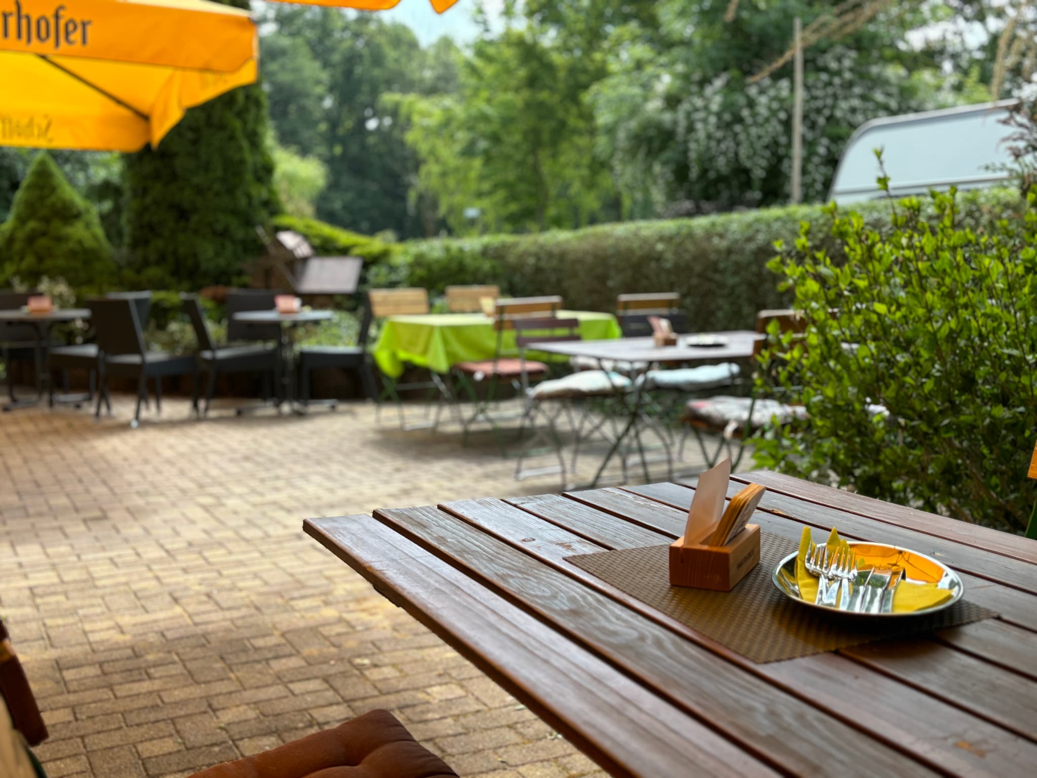 Biergarten mit Holztischen, Stühlen und gelben Sonnenschirmen. Der Sitzbereich ist von Grün umgeben und schafft eine gemütliche Atmosphäre.