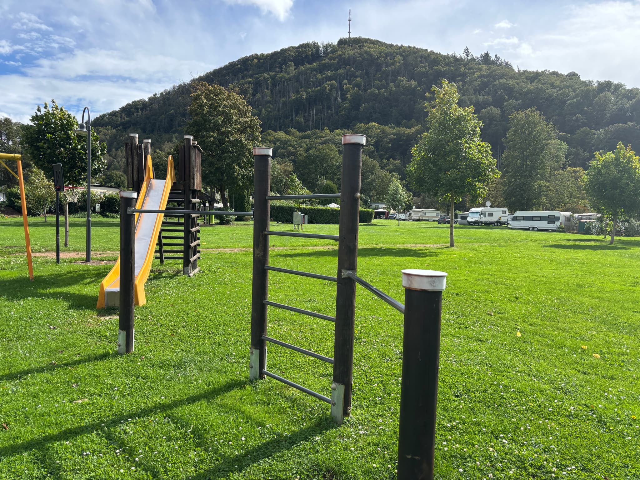 Spielplatz mit Rutsche und Kletterstangen auf einer Wiese, umgeben von Bäumen und Hügeln unter einem teilweise bewölkten Himmel.