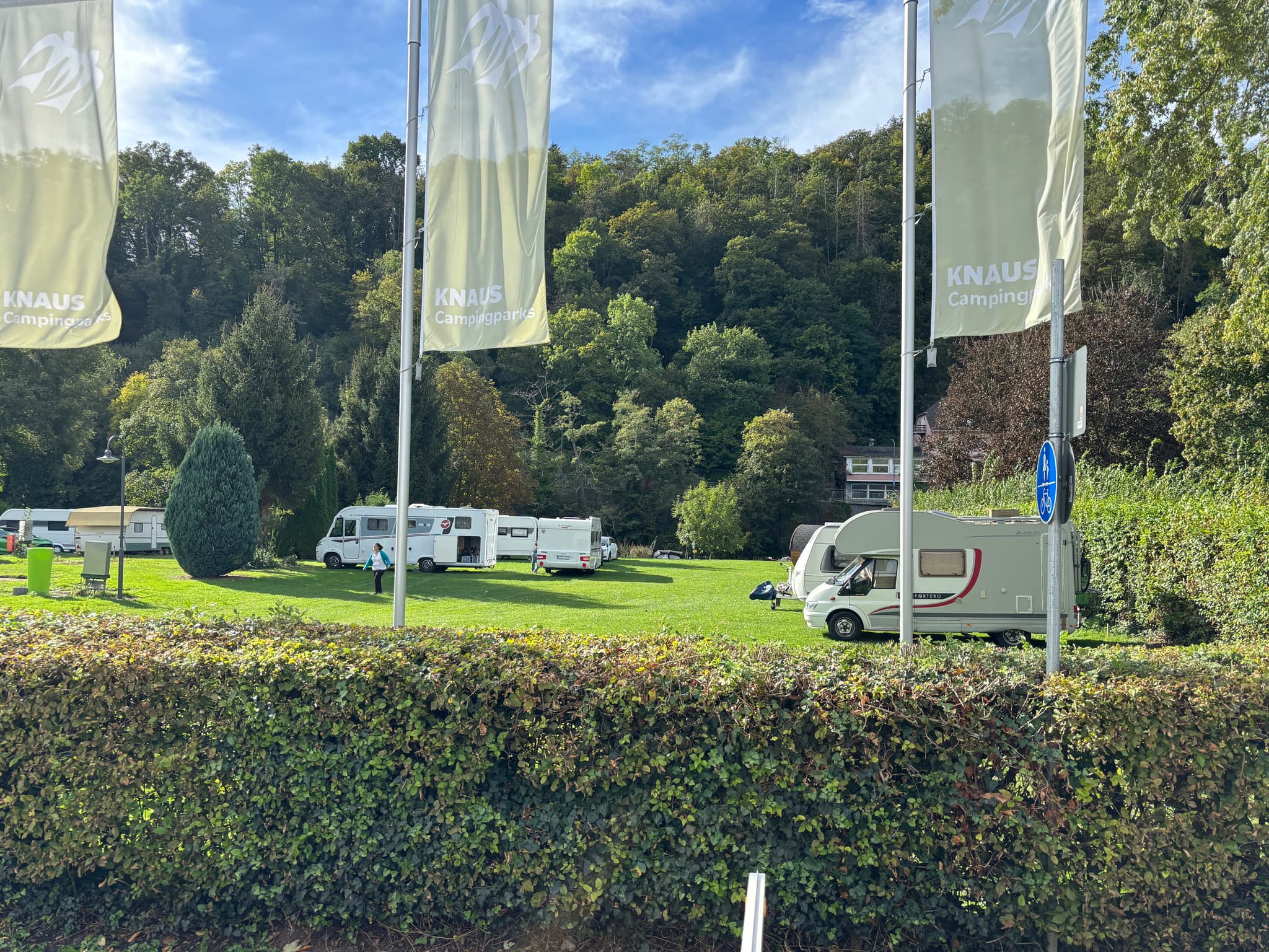 Campingplatz mit mehreren Wohnwagen auf einer Wiese, umgeben von Bäumen und Hügeln unter blauem Himmel, mit hohen Fahnen im Vordergrund.