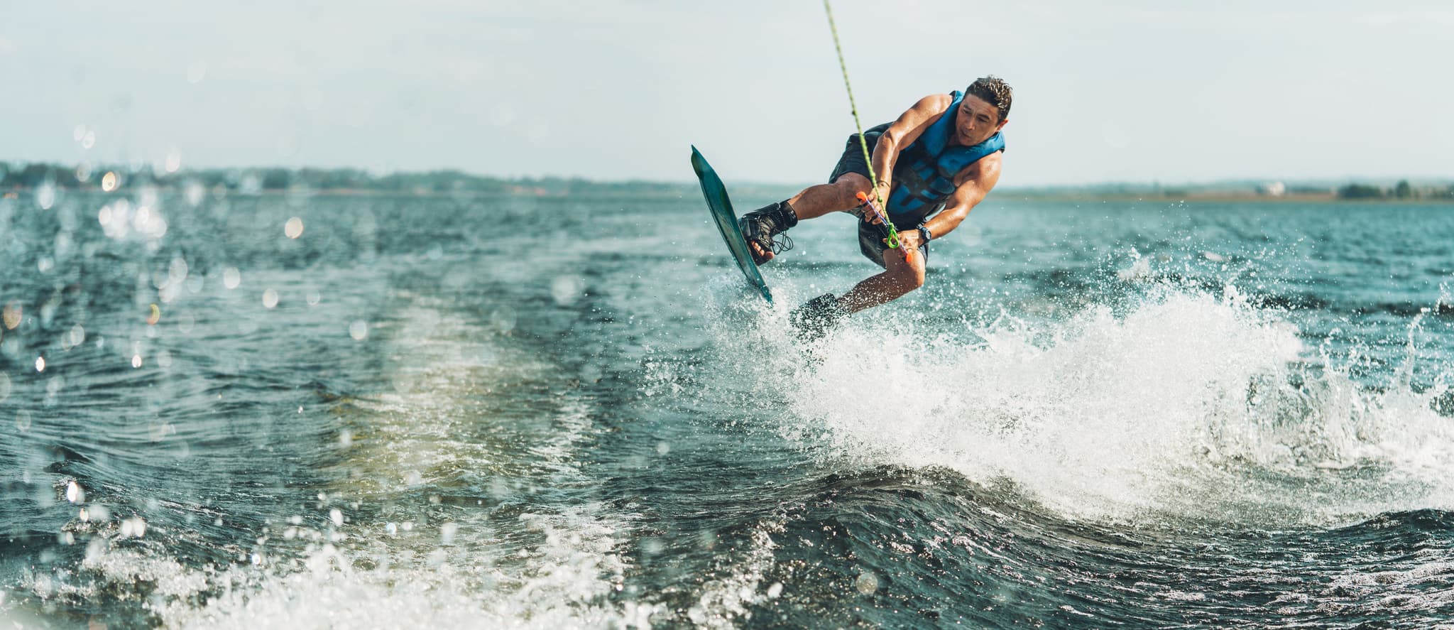 Eine Person in einer blauen Weste springt beim Wakeboarden auf einem See, umgeben von plätscherndem Wasser und unter einem klaren Himmel.