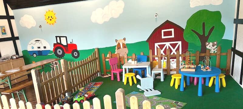 Farbenfroher Kinderspielbereich mit bäuerlicher Wandmalerei, kleinen Tischen, Stühlen, Spielzeug und einem eingezäunten Bereich auf grünem Teppich.