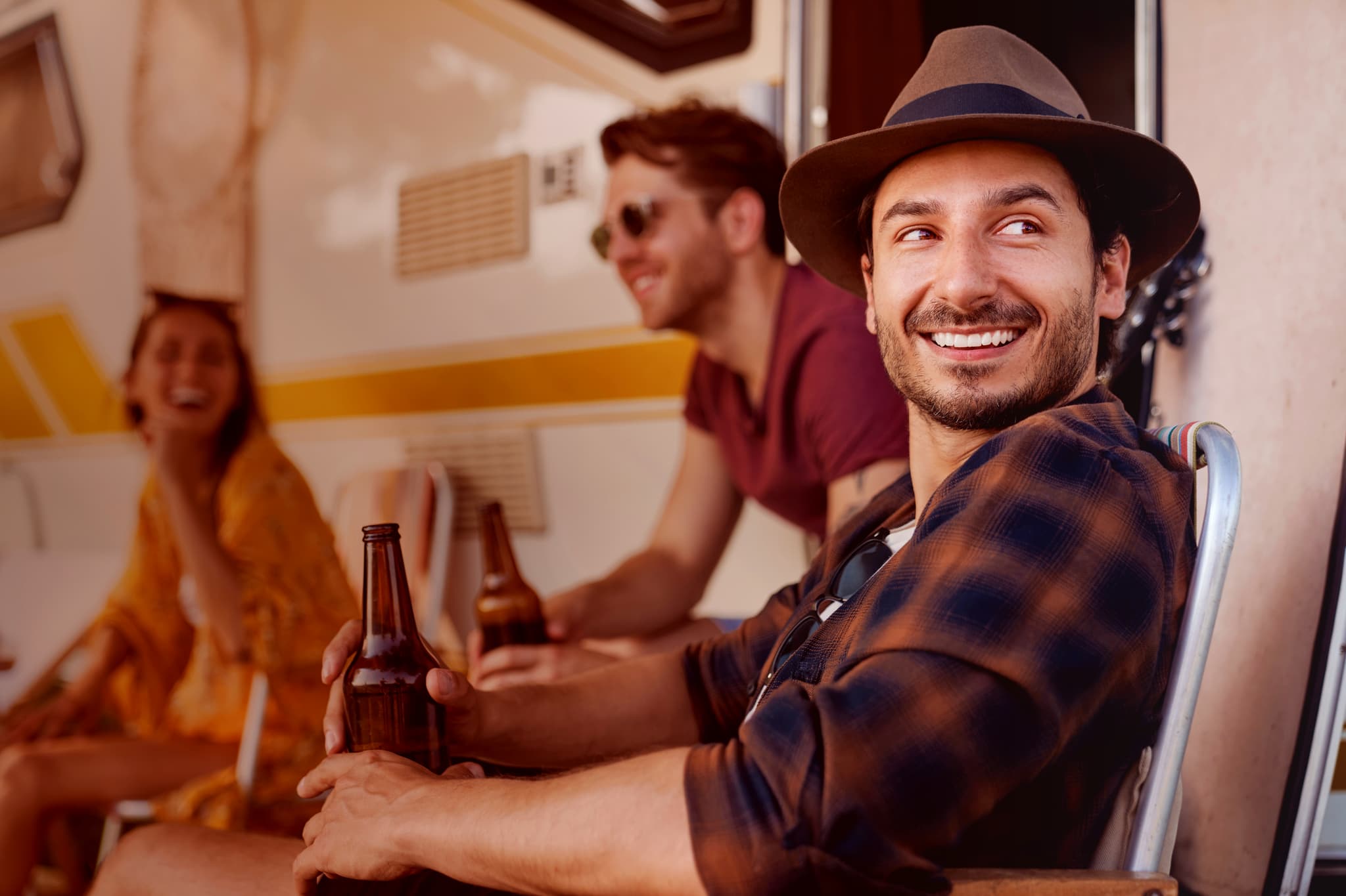Drei Freunde sitzen vor einem Wohnmobil, lächeln und halten Bierflaschen in der Hand. Ein Mann mit einem Hut steht im Vordergrund und schaut zur Seite.