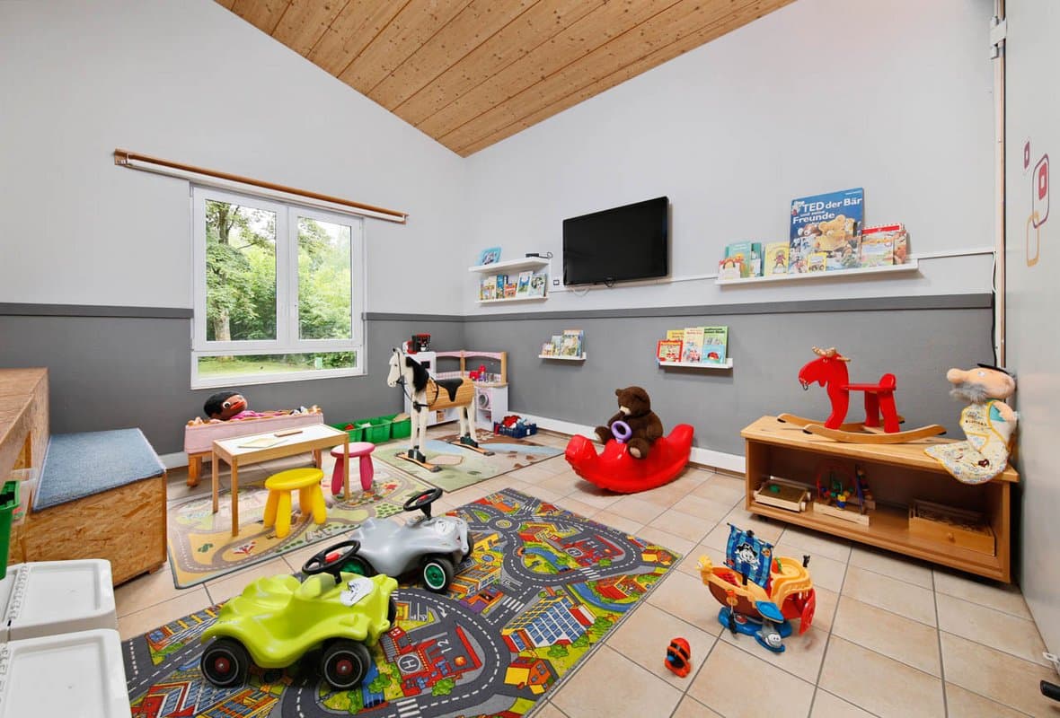 Spielzimmer Ein farbenfrohes Spielzimmer mit Spielzeug, einem kleinen Tisch, Stühlen, einem an der Wand montierten Fernseher und Regalen mit Büchern. Großes Fenster mit Blick ins Grüne.