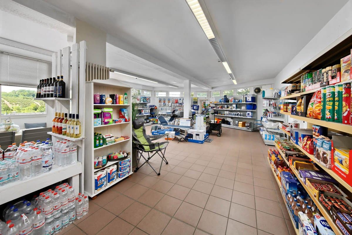 Shop mit frischen Backwaren Ein gut beleuchteter Lebensmittelladen mit Regalen, die mit Getränken, Snacks und Haushaltsartikeln gefüllt sind. Im Gang ist ein Klappstuhl aufgestellt.