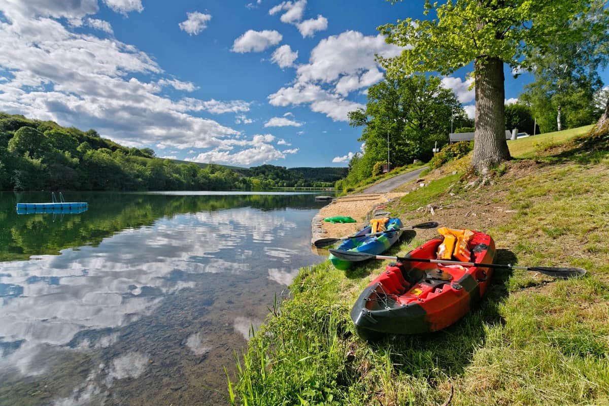 Zwei Kajaks mit Paddeln an einem grasbewachsenen Seeufer unter einem klaren blauen Himmel mit Schäfchenwolken, umgeben von Bäumen und ruhigem Wasser.