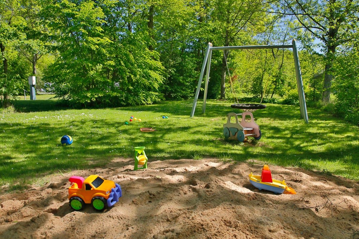 Spielplatz Ein sonniger Spielplatz mit einem Sandkasten, der mit bunten Spielzeugfahrzeugen gefüllt ist. Eine Schaukel und verstreute Spielsachen befinden sich im grasbewachsenen Hintergrund.