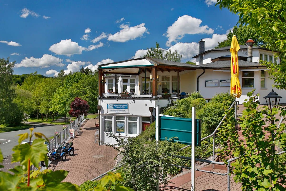 Restaurant mit Terrasse Ein heller, sonniger Tag in einem Restaurant mit Terrasse am Hang, umgeben von üppigem Grün und einem klaren blauen Himmel mit Schäfchenwolken.