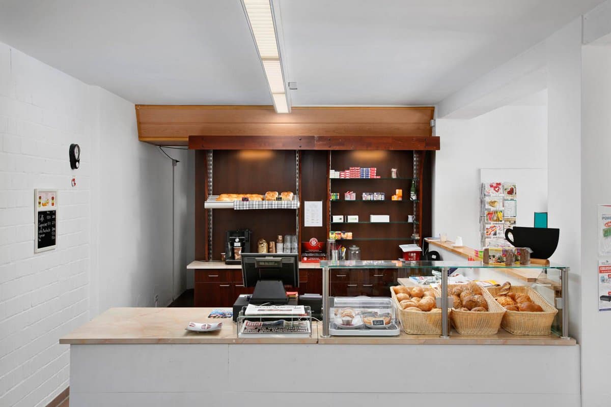 Shop mit frischen Backwaren Eine kleine Bäckertheke mit einer Auslage für Gebäck, Brot und einer Registrierkasse. Dahinter befinden sich Regale mit verschiedenen verpackten Waren und eine Kaffeemaschine.