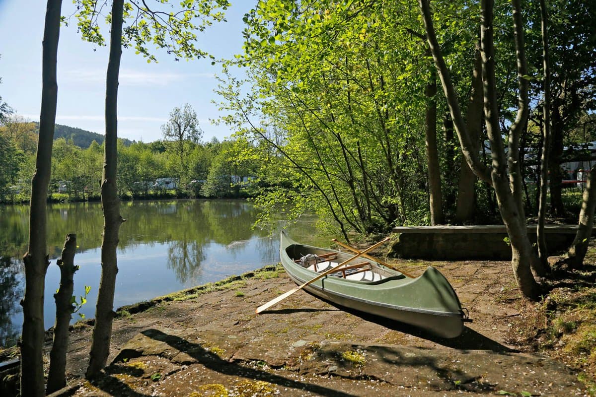 Kanuanlegestelle Ein grünes Kanu liegt an einem sonnenbeschienenen, von Bäumen gesäumten Flussufer mit Rudern darin, umgeben von üppigem Grün und spiegelndem Wasser.