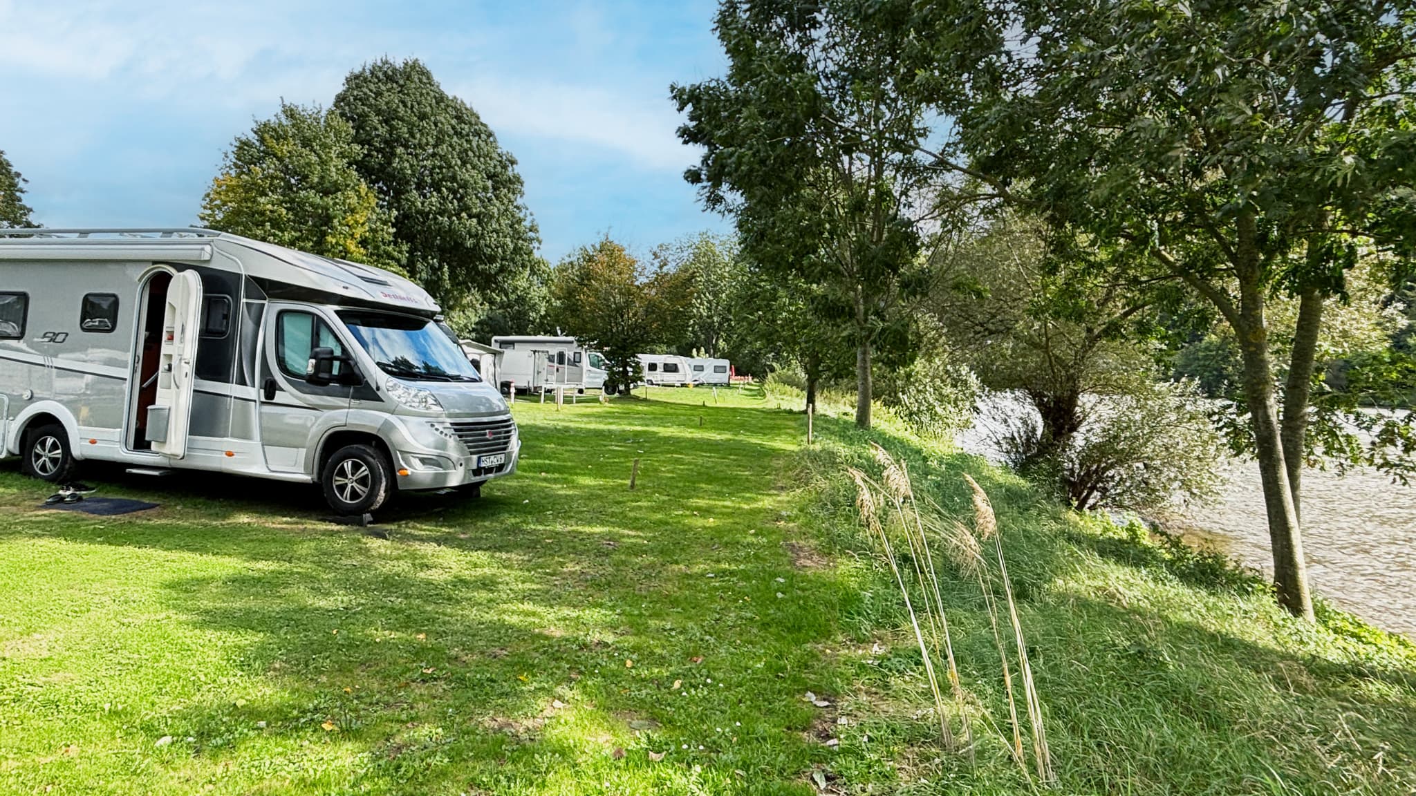 Stellplatz Ufer Ein Wohnmobil, das an einem grasbewachsenen Flussufer geparkt ist, umgeben von Bäumen und anderen Wohnmobilen in der Ferne unter einem klaren blauen Himmel.