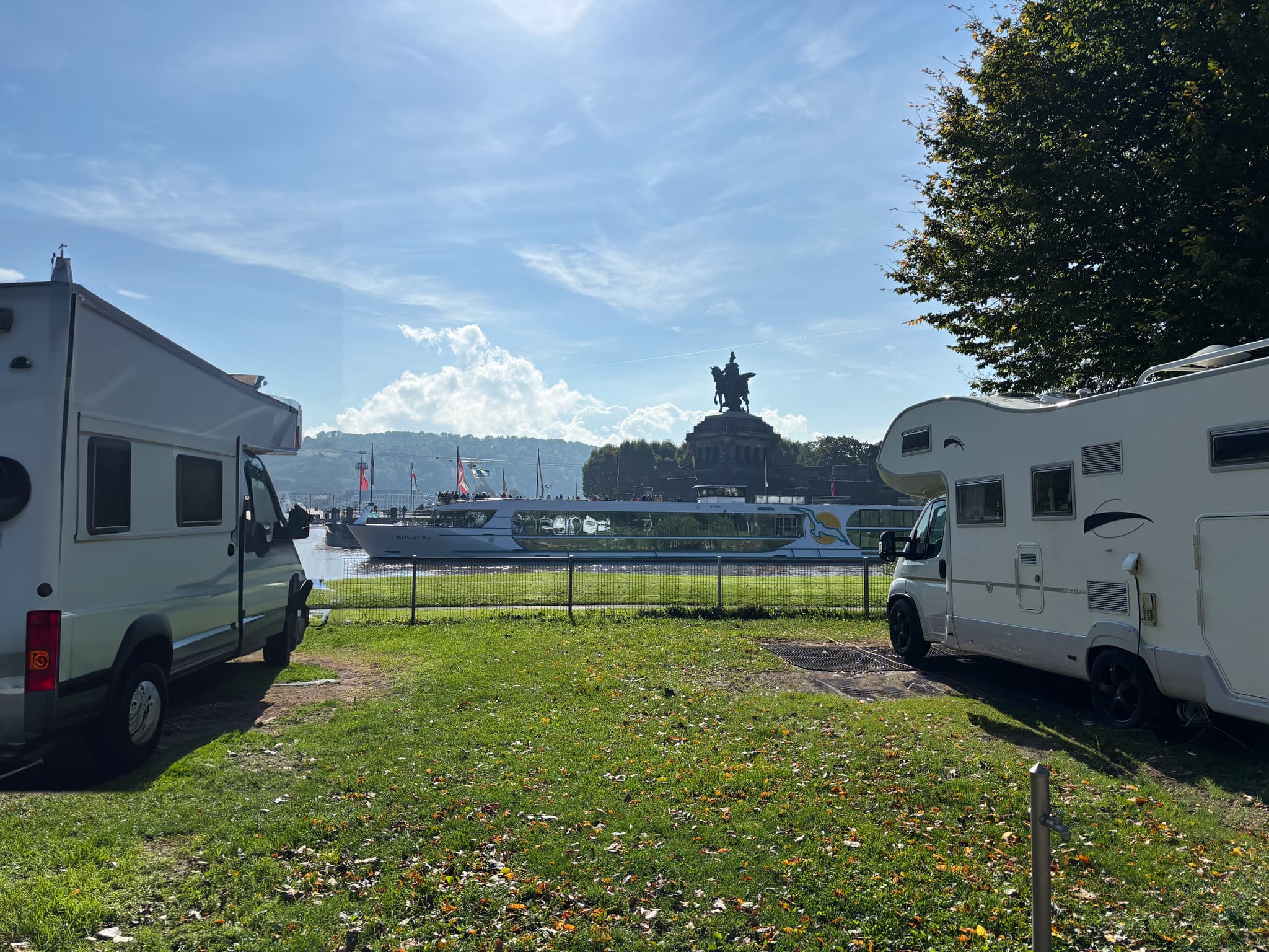 Stellplatz am Wasser Zwei Wohnmobile, die auf einer Wiese in der Nähe eines Flusses geparkt sind, mit einem großen Denkmal und einem Boot im Hintergrund unter einem klaren blauen Himmel.